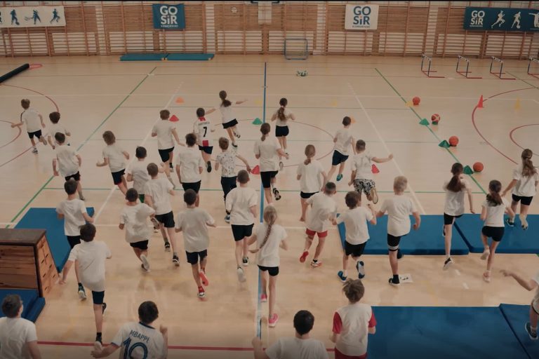 Dzieci wbiegają na szkolną salę gimnastyczną. Stopklatka ze spotu programu edukacyjnego Żyję zdrowo.