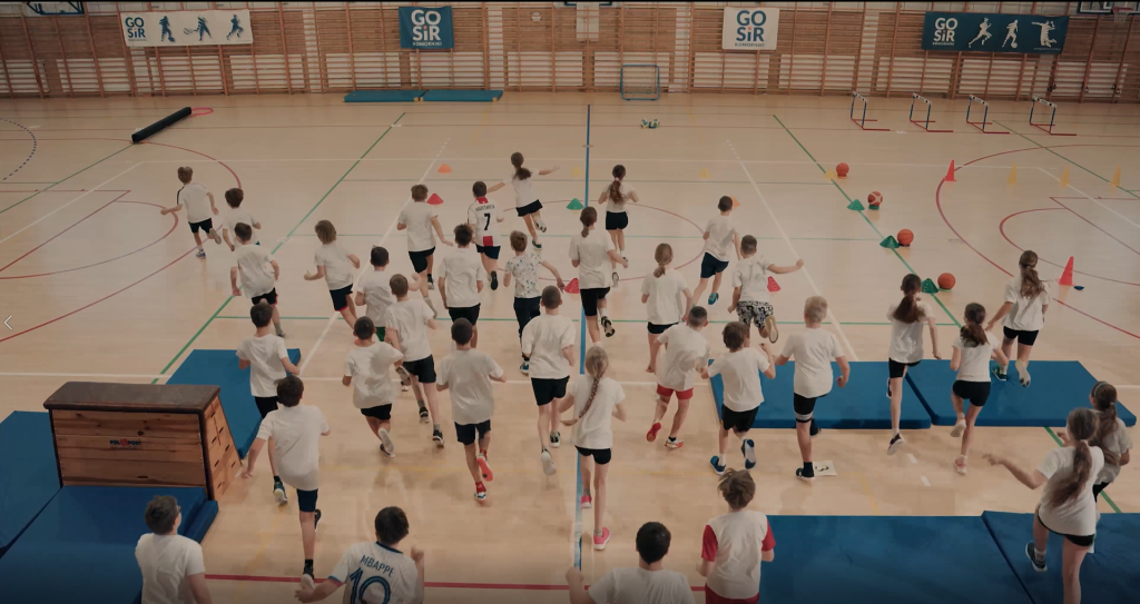 Stopklatka ze spotu "Zawody sportów nietypowych" programu Żyję zdrowo. Scena przedstawiająca dzieci wbiegające na salę gimnastyczną.